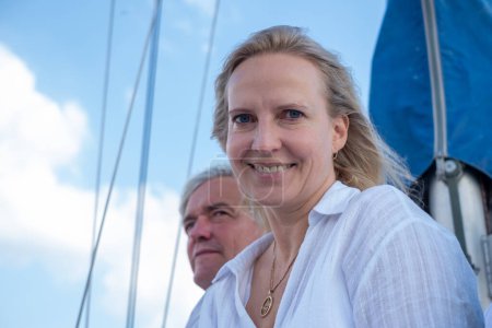 Mujer sonriente en una blusa blanca con un hombre detrás de ella en un velero, compartiendo un momento de alegría bajo un cielo azul, ideal para temas de ocio y viajes. Foto de alta calidad