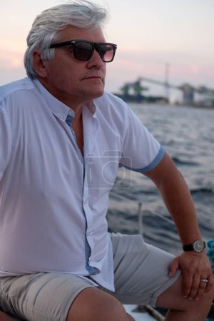 Silver haired man in sunglasses, pensively looking out at the sea from a boat at dusk, capturing a mood of reflection and solitude. High quality photo