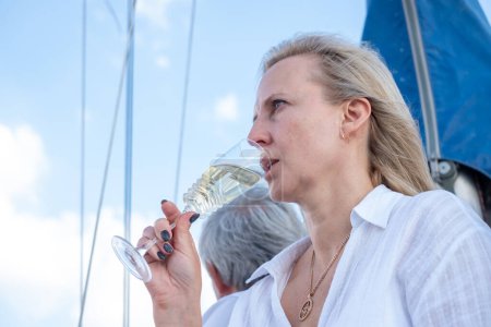 Una escena de yate serena, una mujer rubia disfrutando de una copa de vino, hombre detrás, ambos con vistas al océano, personificando la relajación y la riqueza. Foto de alta calidad