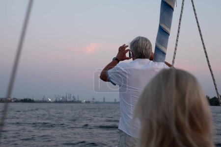 Homme âgé sur un bateau capturant le paysage urbain au crépuscule avec son smartphone, reliant la sérénité de la mer au pouls de la vie urbaine. Photo de haute qualité