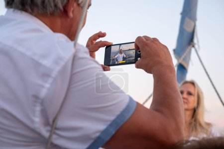 Sobre el hombro vislumbra a un anciano capturando una foto de una mujer en su teléfono, un momento enmarcado por los suaves tonos de un sol poniente. Foto de alta calidad