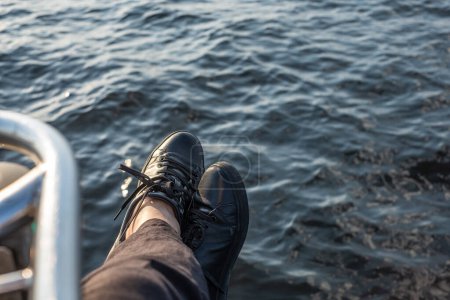 Füße in schwarzen Turnschuhen ruhen auf einem Bootsrand, im Hintergrund das tiefblaue Meer, was einen entspannten maritimen Moment darstellt. Hochwertiges Foto