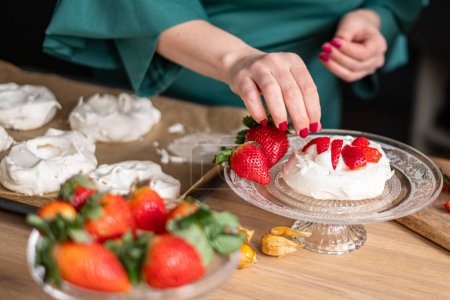 A chef in a green apron assembles strawberry pavlovas in a bright kitchen, with more desserts in soft focus behind. Ideal for culinary websites and dessert blogs. High quality photo