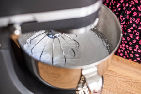Close up of a stand mixer whipping meringue, with a glimpse of a baker in a floral shirt in the background, highlighting the baking process for educational content. High quality photo