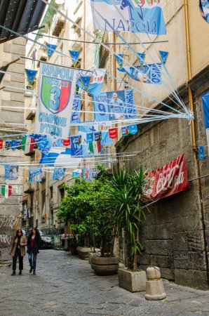 Foto de Nápoles, Italia - 2 de abril de 2023: Dos mujeres caminan a lo largo de la estrecha Strada dell 'Anticaglia, que está ensartada con banderas que celebran el éxito del equipo de fútbol de la ciudad. Nápoles Central. - Imagen libre de derechos