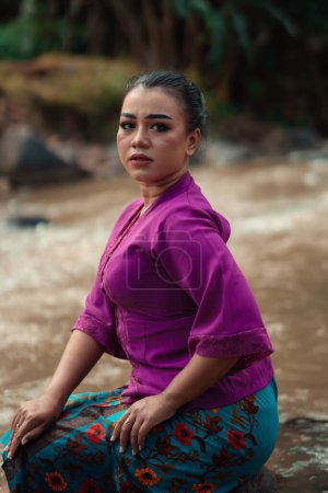Photo for Beautiful Asian woman sitting on the rock near the river with a smiling face while wearing a purple dress inside the village - Royalty Free Image