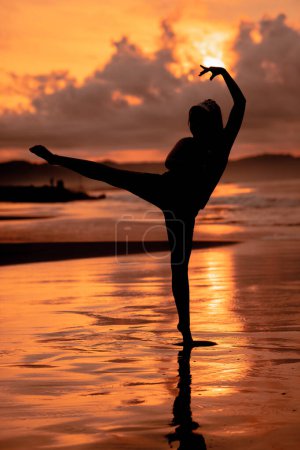 Photo for A Balinese woman in the form of a silhouette performs ballet movements very deftly and flexibly on the beach with the waves crashing in the afternoon - Royalty Free Image