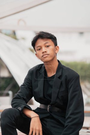 Photo for An Asian teenager in a black denim jacket posing by sitting on a cafe chair and relaxing in the afternoon - Royalty Free Image