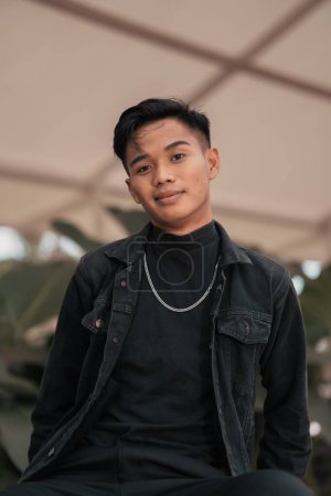 Photo for An Asian man in a black denim jacket and a chain necklace posing in a relaxed style in a park in the afternoon - Royalty Free Image
