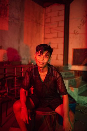 Photo for A gay man wearing black and posing with a chair in an old house with a red light at night - Royalty Free Image