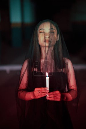 Photo for A female devil worshiper with a transparent veil is performing a spooky ritual by holding a candle in her hand at night - Royalty Free Image