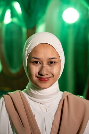 Photo for A Muslim woman with a white headscarf and white clothes smiling very pretty without makeup on her face in a green room at night - Royalty Free Image