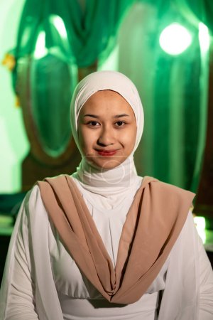 Photo for A Muslim woman with a white headscarf and white clothes smiling very pretty without makeup on her face in a green room at night - Royalty Free Image