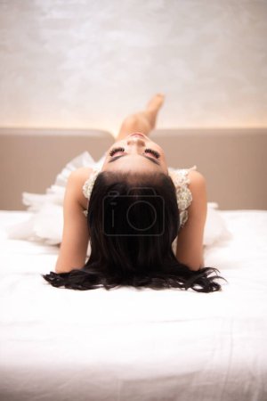 Photo for An Asian woman with black hair is sleeping on a white bed in an elegant hotel at night - Royalty Free Image