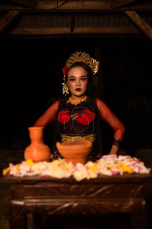 Téléchargez les photos : Une femme asiatique en robe de danseuse rouge est assise à une table d'offrande rituelle pleine de fleurs dans une cabane la nuit - en image libre de droit