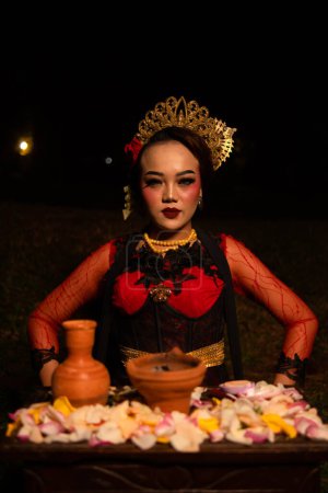 Téléchargez les photos : Une femme asiatique en robe de danseuse rouge est assise à une table d'offrande rituelle pleine de fleurs dans une cabane la nuit - en image libre de droit