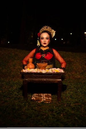 Téléchargez les photos : Plan large d'une fille asiatique en vêtements traditionnels assis dans une forêt avec une table pleine de fleurs la nuit - en image libre de droit