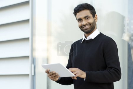 Foto de Handsome smiling Indian man using digital tablet looking at camera standing on the street. Modern technology concept - Imagen libre de derechos