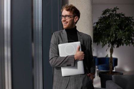 Attractive smiling man wearing eyeglasses holding laptop looking at window