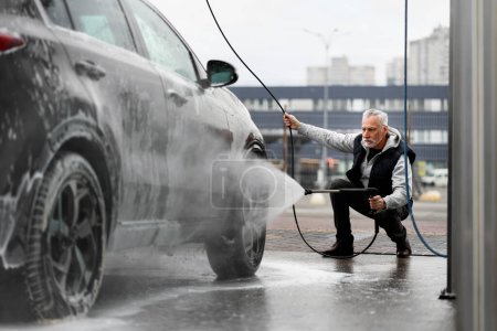 Téléchargez les photos : Elderly man washes his car at touchless self service manual car wash station, using pressurized water hose, spraying water jet on dirty tires while cleaning his dirty vehicle after off road driving - en image libre de droit