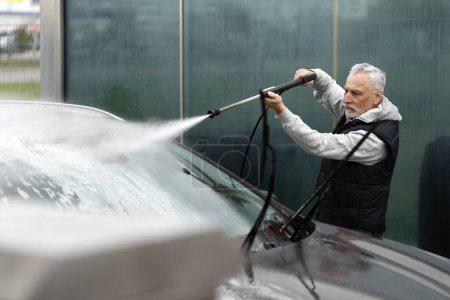 Senior Caucasian white-bearded man, elderly active driver, washing off the detergent foam from his car with water from pressurized hose, at a self-service car wash station. Keeping own vehicle clean