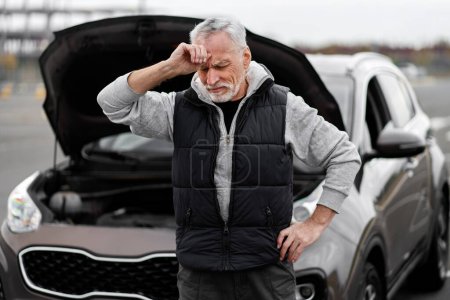 Photo for Perplexed anxious senior man driver, feeling emotional stress, holding hand on head, standing near his broken down auto with open hood, on road. Roadside assistance and car insurance. Engine failure - Royalty Free Image
