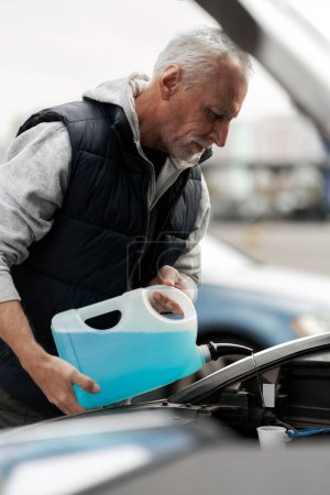 Foto de Close-up elderly man, 60-70 years driver, standing by his land vehicle with open hood, filling the reservoir with blue fluid windscreen washer. The concept of car maintenance - Imagen libre de derechos