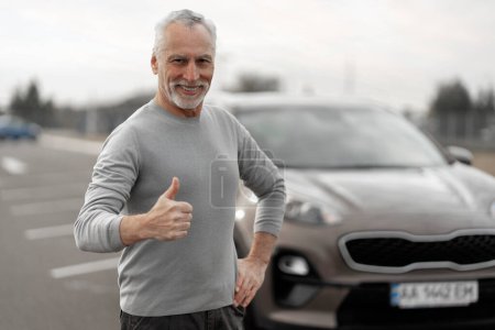 Téléchargez les photos : Handsome gray-haired senior man in casual clothing, expressing positive emotions, showing thumb up, smiling a cheerful toothy smile, standing near his modern comfortable car on the automobile parking - en image libre de droit