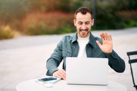 Photo for Young attractive smiling man using laptop computer waving hand having video call. Happy successful influencer recording video sitting in park. Modern technology, video conference concept - Royalty Free Image