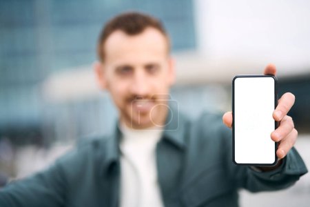 Photo for Smiling modern man holding mobile phone showing empty display, selective focus. Hipster male using mobile app. Mockup - Royalty Free Image
