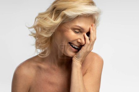 Téléchargez les photos : Delightful blonde, happy mature woman with fresh clean skin, laughing while covering her face with her hand, isolated on white background. Skin and body care concept. Menopause. Copy advertising space - en image libre de droit