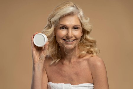 Téléchargez les photos : Close-up portrait of Caucasian attractive blonde woman, holding a jar with anti-aging rejuvenating moisturizing beauty product, smiling a toothy smile looking at camera, isolated over beige background - en image libre de droit