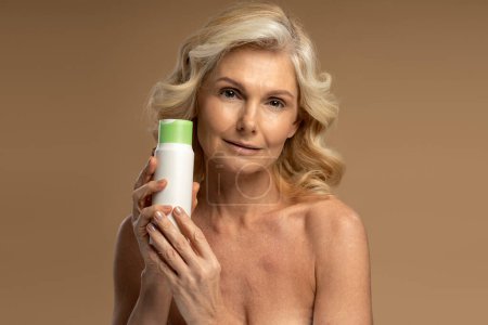 Téléchargez les photos : Confident beautiful Caucasian shirtless, 50s mature woman looking at camera, holding a jar with anti-aging smoothing moisturizing cosmetic product, isolated beige background. Mature beauty concept - en image libre de droit