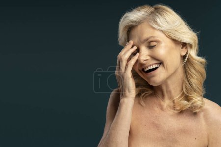 Téléchargez les photos : Delightful Caucasian shirtless blonde mature woman, with her eyes closed, smiling a beautiful toothy smile, touching her face, laughing, isolated over dark colored background. Skin care concept - en image libre de droit