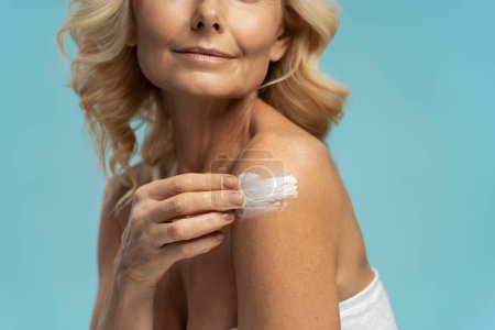 Téléchargez les photos : Close-up of a Caucasian mature woman applying anti-aging smoothing moisturizer with uplift effect, on her body, isolated on blue background. Spa procedure. Skin and body care. Mature beauty concept - en image libre de droit