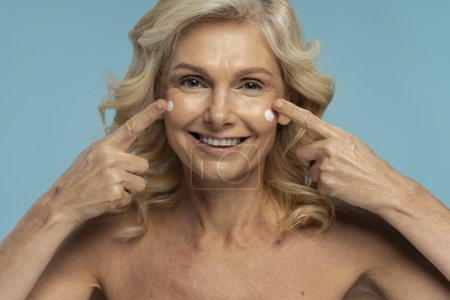 Foto de Gorgeous 50s Caucasian blonde senior woman, smiling a toothy smile looking at camera, applying anti-aging smoothing moisturizing face cream, isolated over blue background. Mature beauty concept - Imagen libre de derechos