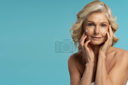Téléchargez les photos : Confident portrait of Caucasian mature woman, applying anti-aging face cream, holding hands on her face and confidently looking at camera, posing shirtless over blue background with copy ad space - en image libre de droit