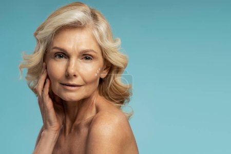 Téléchargez les photos : Beautiful 50-55 years old Caucasian blonde mature woman applying anti-aging moisturizing cream onto face, isolated on blue background with copy advertising space. Skin care and mature beauty concept - en image libre de droit