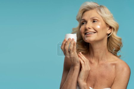 Téléchargez les photos : Confident attractive Caucasian shirtless, 50s mature woman smiling, holding a jar with anti-aging smoothing moisturizing cosmetic product, isolated on blue colored background. Mature beauty concept - en image libre de droit