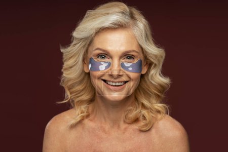 Téléchargez les photos : Beautiful Caucasian mature woman with hydrogel collagen eye patches, smoothing dark circles and puffiness under the eyes smiling looking at camera, posing shirtless on color background. Skin care - en image libre de droit