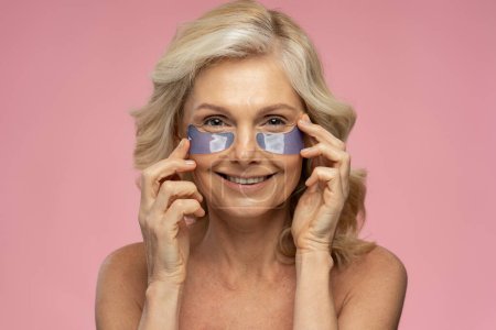 Téléchargez les photos : Attractive mature woman with half-naked body and no makeup, touching face and smiling at camera while using under-eye patches for dark circles removal, making skin fresh and glowing, pink background - en image libre de droit
