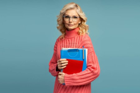 Charming confident Caucasian mature woman with blond wavy hair, wearing eyeglasses and dressed in warm pink sweater, posing with notepads and textbooks, looking at camera isolated on blue background