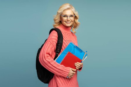 Téléchargez les photos : Successful 50-55 years old mature blonde woman, a confident school teacher carrying a backpack and notepads, cutely smiling looking at camera, isolated on blue colored background. Education concept - en image libre de droit