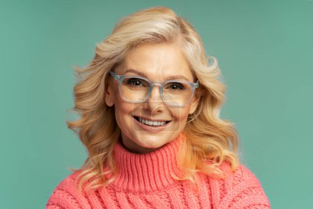 Téléchargez les photos : Isolated close-up portrait on colored background of a confident charming Caucasian mature woman with beautiful wavy blond hair, wearing eyeglasses, smiling a beautiful toothy smile, looking at camera - en image libre de droit