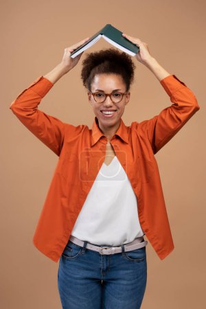 Photo for Waist-up portrait of a confident African American young woman in bright orange shirt and blue jeans, smiling at camera, holding a book like a house roof above her head, isolated over beige background - Royalty Free Image