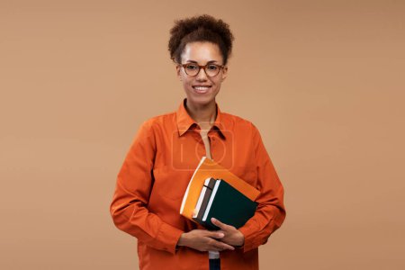 Photo for Confident young African American woman, high school teacher, tutor wearing glasses and orange casual shirt, posing with books, isolated on beige color background. College, Knowledge, Education concept - Royalty Free Image