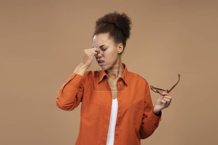 Foto de Overworked multitasking African American business woman, sales manager holding glasses, massaging her nose bridge, feeling headache after hardworking day in office, isolated beige background. Ad space - Imagen libre de derechos