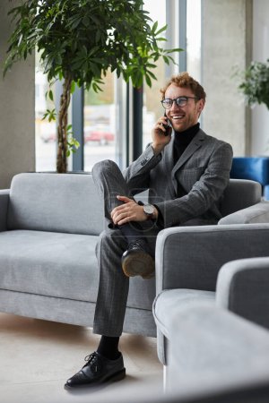 Téléchargez les photos : Confident businessman in gray formal suit and eyeglasses, smiles while talks on mobile phone, sitting on a comfortable sofa in the VIP lounge of a modern office interior. People Business Communication - en image libre de droit