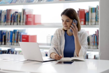 Foto de Joven mujer de negocios pensativa hablando en el teléfono móvil, utilizando el ordenador portátil y tomando notas sentado en la oficina moderna. Multitarea, concepto de tecnología - Imagen libre de derechos