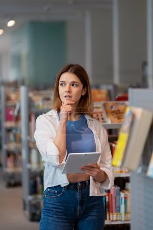 Foto de Joven mujer pensativa sosteniendo tableta digital mirando hacia otro lado de pie en la tienda de libros - Imagen libre de derechos
